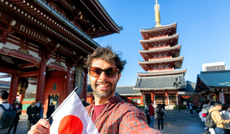 Handsome young tourist enjoying summer holiday in Tokyo, Japan - Traveling life style concept with smiling man taking selfie on city street with japan flag-