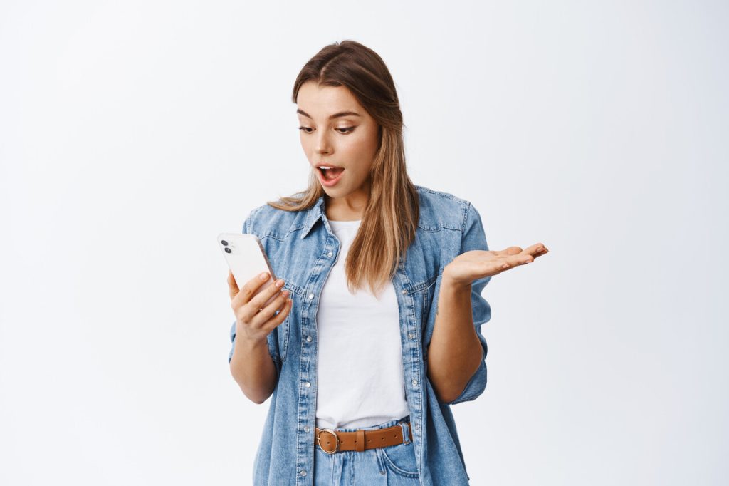 excited woman gasping fascinated while reading message on mobile phone. Girl checking smartphone, looking at screen with pleased face, white background